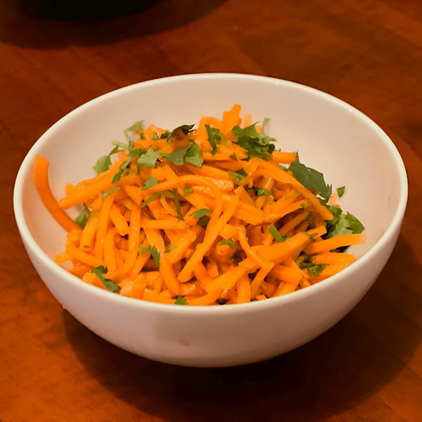 A white bowl filled with bright orange shredded carrots and green herbs on a wooden table.