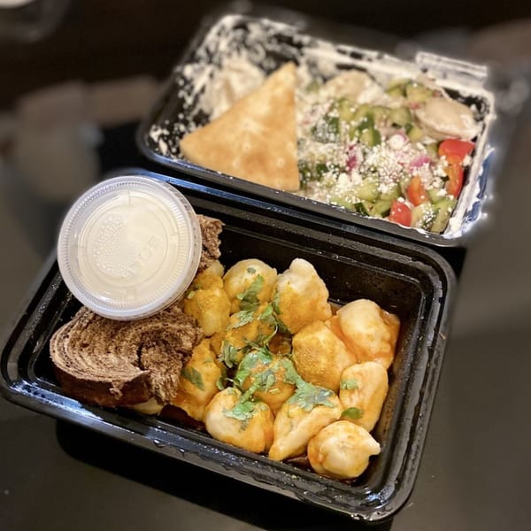 Two takeout containers with Pel Meni dumplings, bread, sauce, and a salad with pita.